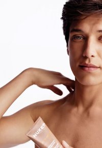 NUXE-produkt i en tub som hålls i handen, mjuk beige med vit text, bredvid en bar axel; slät hud, ljus bakgrund.