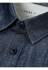 Ciemnoniebieska koszula z denimu o fakturowanym wykończeniu, z ostrym kołnierzykiem i czarnymi guzikami. Etykieta widoczna wewnątrz kołnierza.