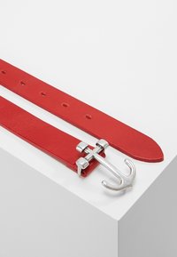 Ceinture en cuir rouge avec une boucle en métal argenté en forme d'ancre reposant sur une surface blanche.