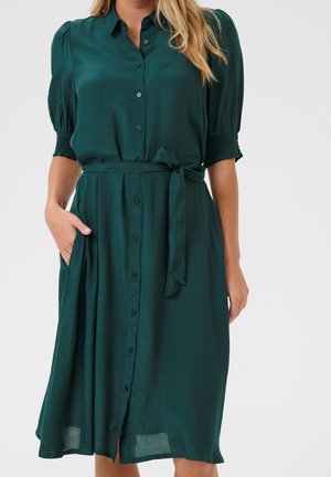 Femme portant une robe midi boutonnée bleu sarcelle avec des manches bouffantes, des poignets smockés, une ceinture à la taille et la main dans la poche, sur un fond uni.