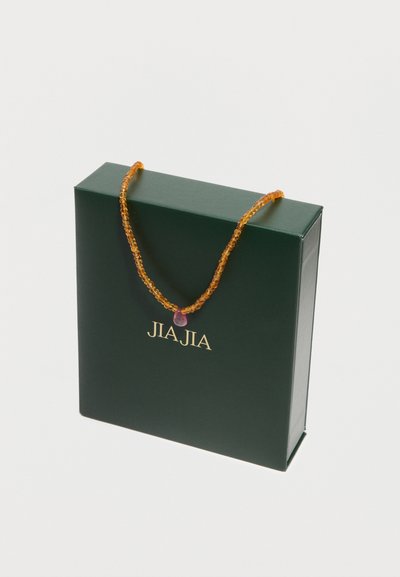 Boîte de marque JIA JIA vert foncé avec un collier de perles ambrées présentant un pendentif unique en forme de goutte rose, posé dessus.