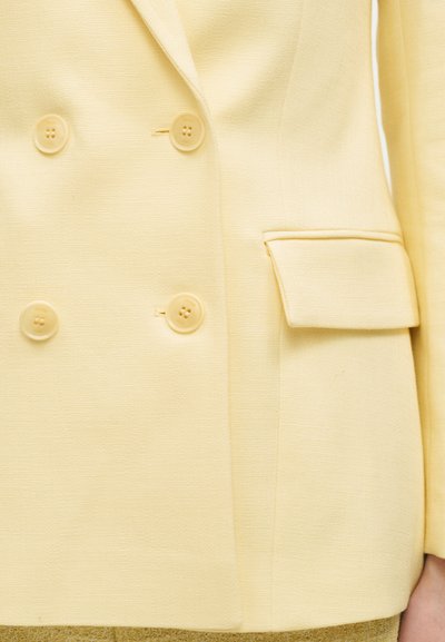 Blazer double boutonnage jaune clair avec quatre boutons et une poche à rabat côté droit, porté par une personne avec le bord de la manche visible.
