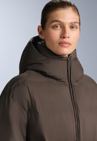 Hnedá bomber bunda s vysokým golierom a zipsom. Hladká, polstrovaná textúra s minimalistickým dizajnom a žiadnymi výraznými vzormi.