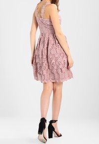 Höyhenen vaaleanpunainen pitsimekko kukkakuviolla, vartalonmyötäinen yläosa, levenevä helma ja säädettävät olkaimet. Yhdistetty mustiin nilkkaremmi-korkokenkiin.