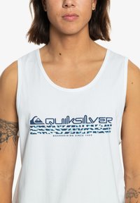 Valkoinen tanktoppi, jossa on sininen ja teallogo graafisella kuviolla raitojen kanssa, "Quiksilver"-teksti ja sileä puuvillapinta.