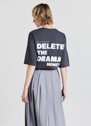 T-shirt oversized grigio scuro con testo bianco sulla schiena che recita "CANCELLA IL DRAMA HONEY", abbinato a una gonna grigia plissettata fluida.
