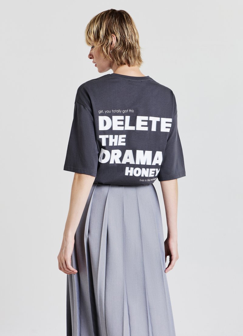 T-shirt oversized grigio scuro con testo bianco sulla schiena che recita "CANCELLA IL DRAMA HONEY", abbinato a una gonna grigia plissettata fluida.