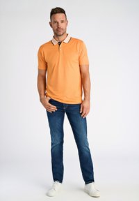 Pomarańczowa koszulka polo z czarnym i beżowym kołnierzykiem, w połączeniu z niebieskimi dżinsami i białymi trampkami, stojąca na jednolitym tle.