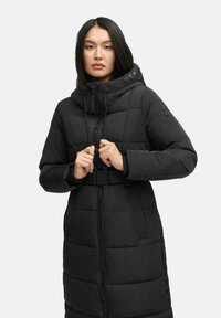 Manteau long noir matelassé avec capuche, doté d'une fermeture éclair, de cordons de serrage ajustables et d'une taille ceinturée. Des panneaux texturés ajoutent des détails.