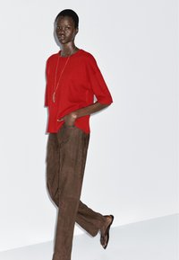 Czerwony sweter z dzianiny, krótki rękaw i ściągacz na dole, w połączeniu z brązowymi zamszowymi spodniami o szerokim kroju i sandałami, dopełniony naszyjnikiem z wisiorkiem.