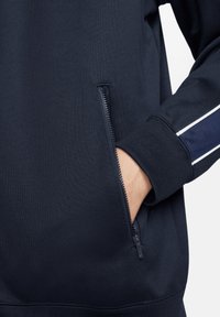 Sudadera de marina con un bolsillo con cremallera y detalles de franjas blancas en contraste en la manga. Tejido suave y texturizado con puños acanalados.