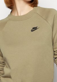 Close-up of a person wearing an olive-green Nike sweatshirt with a small black Nike logo on the upper chest.