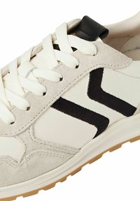 Weiß-beige Sneaker mit Obermaterial aus Wildleder und Leder, schwarzen Akzenten und einer strukturierten Gummisohle mit einem ausgeprägten Chevron-Muster.