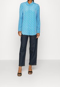 Camicia blu a maniche lunghe con motivo bianco ondulato, colletto con bottoni, abbinata a pantaloni larghi in denim scuro e tacchi neri open-toe.
