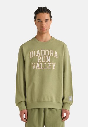 Giovane uomo con capelli intrecciati che indossa una felpa verde oliva con la scritta "DIADORA RUN VALLEY" e pantaloni coordinati su sfondo semplice.