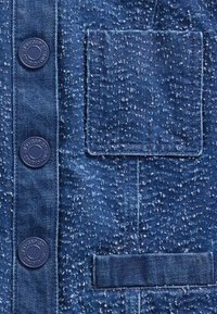 Jeans tkanina v temno modri barvi z teksturirano površino. Vsebuje dva velika modra gumba in zakrpani žep z vodoravno vrstico pod njim.