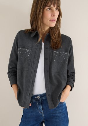 Grå skjorte med knapper og to brystlommer, udsmykket med dekorative rhinsten. Båret over en hvid top, sat sammen med blå denim jeans.