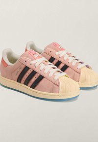 Jasnoróżowe zamszowe trampki z trzema czarnymi paskami, kremowym gumowym noskiem oraz kolorową etykietą z postacią na języku.