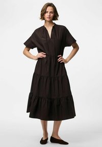 Melna slāņota kleita ar īsām piedurknēm, sagatavotu kakla izgriezumu un plūstošu apakšmalu. Izgatavota no viegla auduma, saskaņota ar brūnām kurpēm.