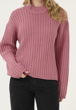 Woman wearing a dusty pink ribbed knit sweater with long sleeves and black jeans, standing against a neutral background.