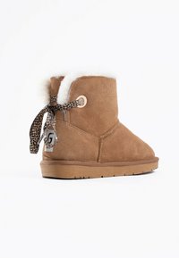Botín de tobillo de ante color beige con un forro de piel sintética blanca, cinta de estampado de leopardo y un acento con logo redondo. Suela plana y texturizada para agarre y comodidad.