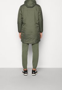 Parka imbottito verde con cappuccio, maniche lunghe e orlo curvo; abbinato a pantaloni jogger verdi coordinati e scarpe sportive nere.