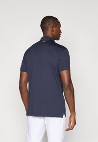 Námořnická modrá polo tričko, krátké rukávy, žebrovaný límec, boční rozparky, hladká textura, detail loga na zadní části límce. Nosené s bílými kalhotami.