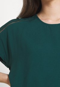 Haut à manches courtes bleu sarcelle avec un col rond, une couture texturée subtile le long des épaules, et une coupe décontractée dans un tissu lisse.