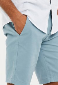 Světle modré bavlněné šortky s bočními kapsami, s hladkým povrchem a přizpůsobeným střihem, v kombinaci s bílou lněnou košilí.