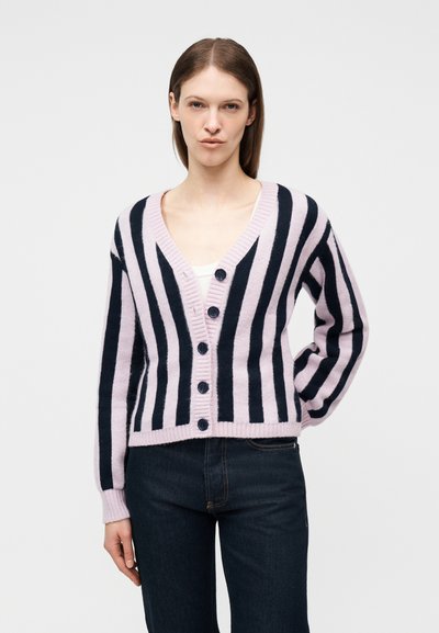 Γυναίκα με καστανά ίσια μαλλιά φοράει ένα ροζ και μαύρο κατακόρυφα ριγωτό ζακέτα με κουμπιά και σκούρο τζιν, στέκεται μπροστά σε λευκό φόντο.