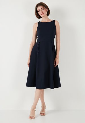 Kobieta ubrana w granatową, bezrękawnikową sukienkę midi z dopasowanym gorsetem i rozkloszowanym dołem, stojąca na tle jednolitego białego tła.