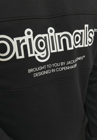 Sudadera negra con gran texto blanco "Originales" y texto blanco más pequeño debajo, hecha de una tela suave con un diseño clásico de cuello redondo.
