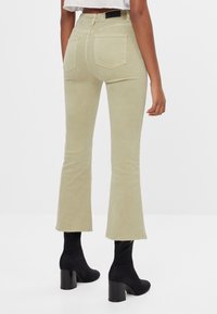 Jeans de campana de color verde claro con bajos deshilachados. Cuenta con dos bolsillos traseros y una etiqueta negra en la cinturilla. Combinados con botas de tacón negras.