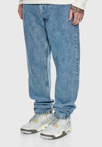 Light blue baggy jeans worn with white and gray sneakers, partial view of tattooed hand and white long-sleeve top on gray background.