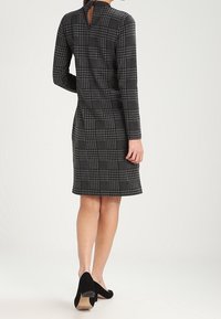 Robe à carreaux noir et gris avec des manches longues, un détail en forme de trou de serrure au dos, et une silhouette ajustée, associée à des chaussures à talons hauts noires.