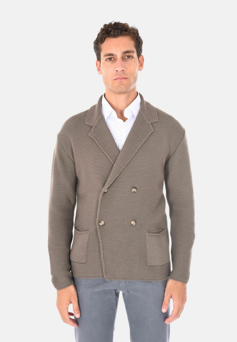 Giacca marrone strutturata doppiopetto con rever a punta e due tasche frontali, indossata sopra una camicia bianca.