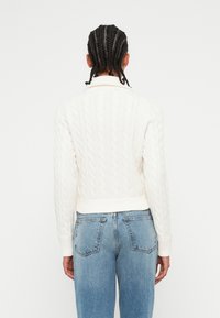 Pull en maille torsadée crème avec un design raccourci et des poignets côtelés, associé à un jean bleu clair ample.