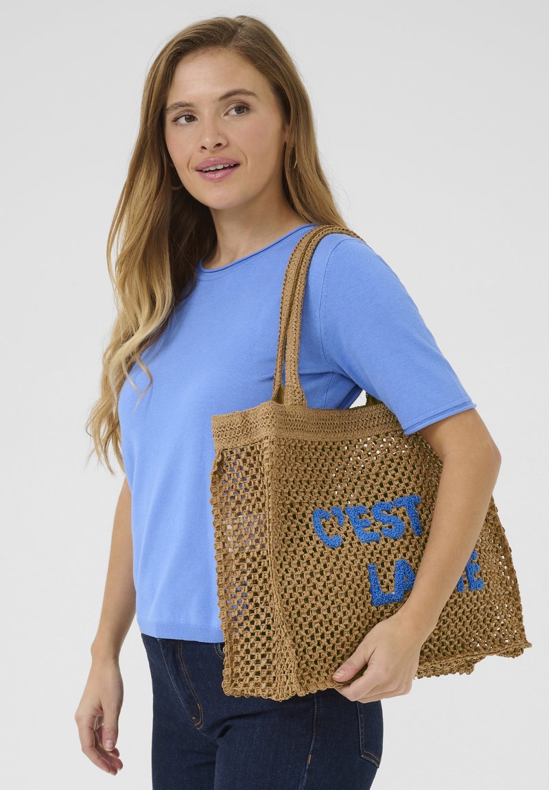 Brun virkad tote-väska med blått "C'EST LA VIE"-text, med en rymlig öppen design och stadiga axelremmar, tillsammans med en blå skjorta.
