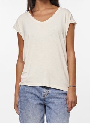 Vrouw met een beige, geribd T-shirt met korte mouwen en lichtblauwe spijkerbroek, staand tegen een effen witte achtergrond.