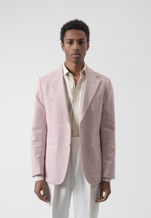 Joven con blazer de cuadros rosa claro sobre una camisa crema y pantalones blancos, posando frente a un fondo gris claro liso.