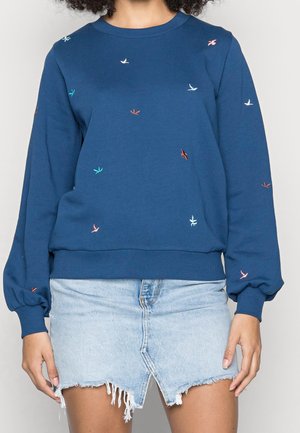 Mujer con sudadera azul con pequeños patrones de pájaros coloridos y una minifalda vaquera desgastada azul claro.
