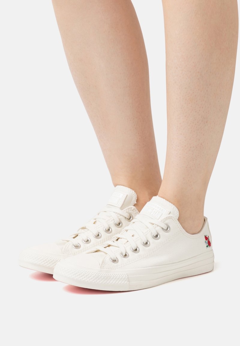 Paire de baskets basses blanches avec lacets et petite broderie de fleur rouge sur le côté extérieur, portées pieds nus sur fond blanc.