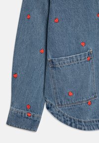 Denimskjorte med rød hjertebrodering, rund kant og brystlomme. Stoffet er blått med en subtil tekstur.