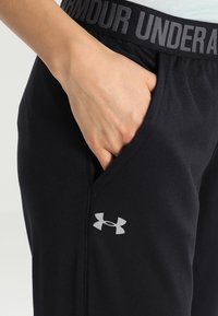 Svarta träningsbyxor med ett grått elastiskt midjeband med texten "UNDER ARMOUR". Inkluderar sidofickor och en liten grå logotyp.