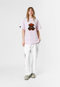 Camiseta de algodón rosa claro con un gráfico de oso de peluche, combinada con pantalones blancos de corte relajado que tienen cremalleras y paneles laterales negros.