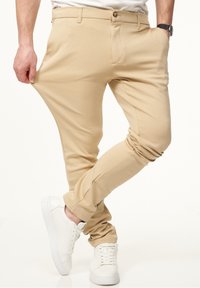 Beige Stretchhosen mit glatter Textur, ausgestattet mit einem Knopfverschluss und seitlichen Taschen, kombiniert mit weißen Sneakers.