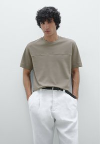 Camicetta khaki a maniche corte con una texture liscia, caratterizzata da una cucitura orizzontale sul petto, abbinata a pantaloni bianchi a vita alta.