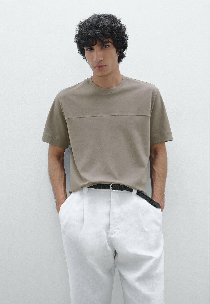 Camicetta khaki a maniche corte con una texture liscia, caratterizzata da una cucitura orizzontale sul petto, abbinata a pantaloni bianchi a vita alta.