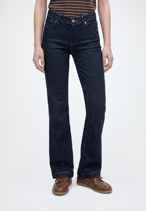 Personne portant un jean bootcut bleu foncé, des chaussures bateau marron et une chemise rayée marron, debout devant un fond clair uni.