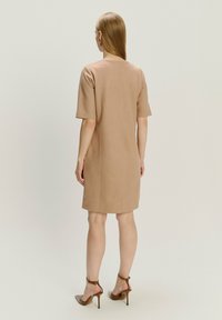 Frau mit glattem roten Haar, die ein knielanges beiges Kleid und braune High Heels trägt, steht mit dem Rücken zu einer einfarbigen Hintergrund.